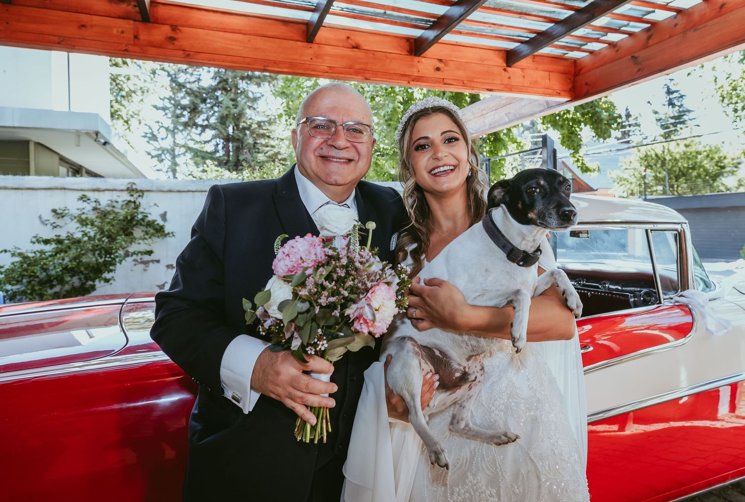 Smiling bride in a white gown poses with an older man holding flowers and a dog by a red car.