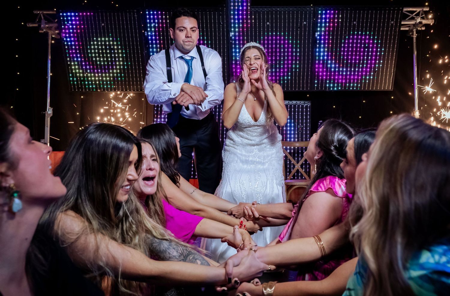 Bride reacts with joy as wedding guests eagerly reach toward her on a colorful dance floor.