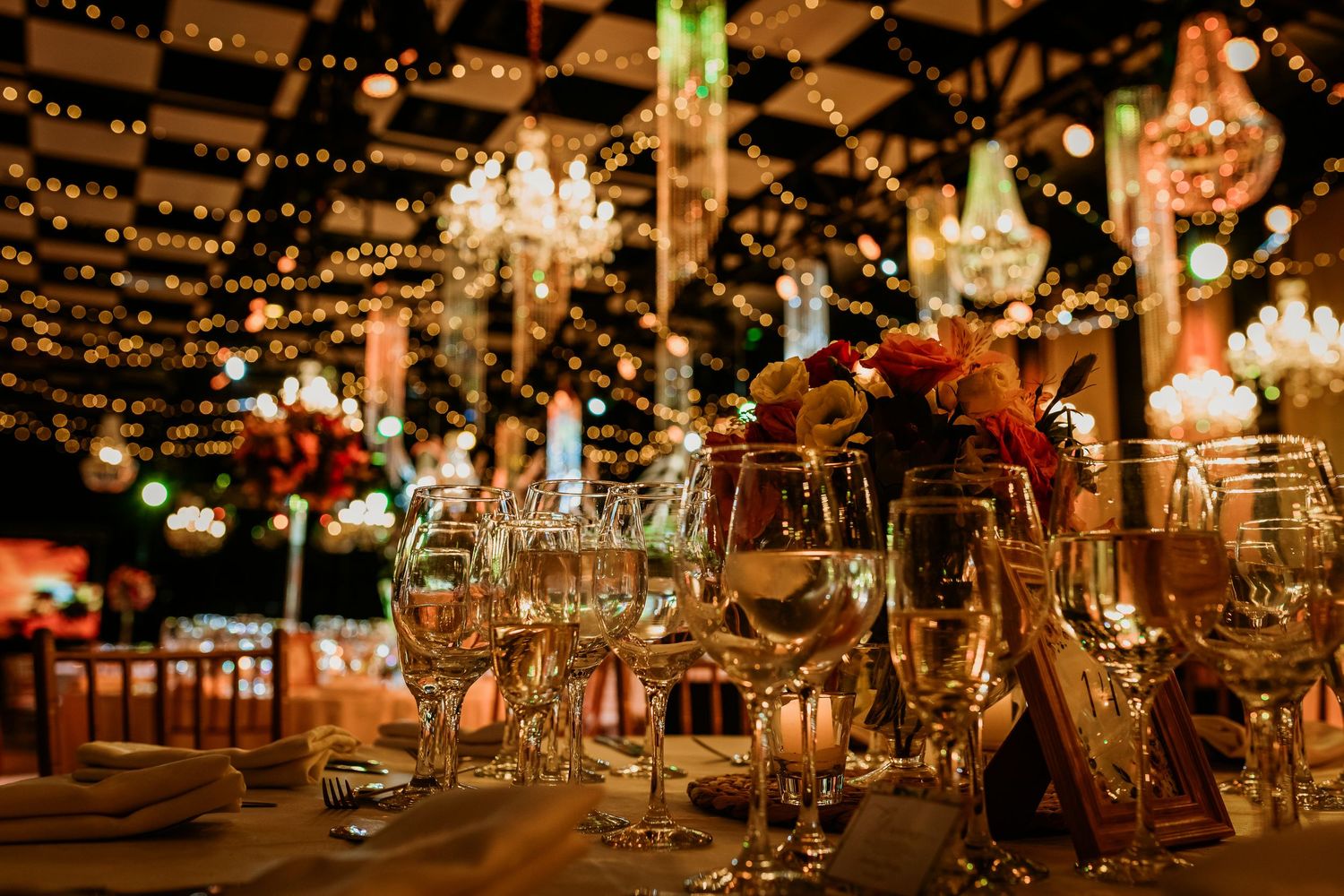 Elegant wedding reception table setting with crystal wine glasses, string lights, and chandelier ambiance.