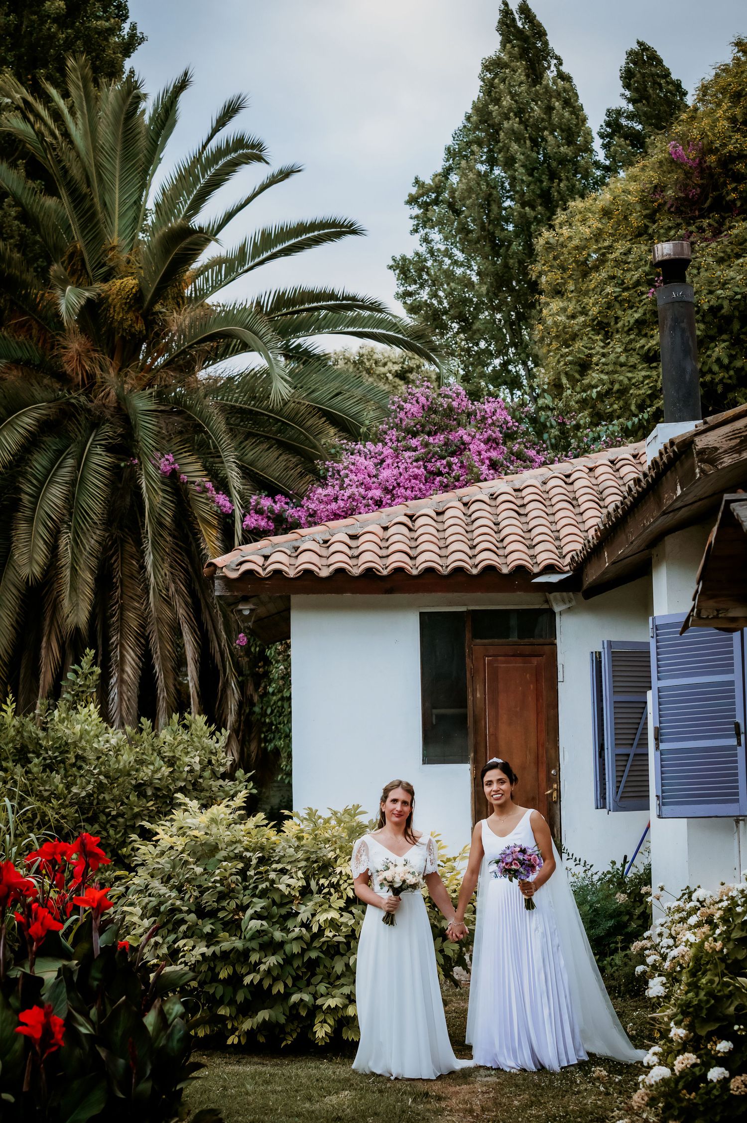 Two brides in white gowns hold bouquets outside a charming cottage surrounded by tropical gardens.