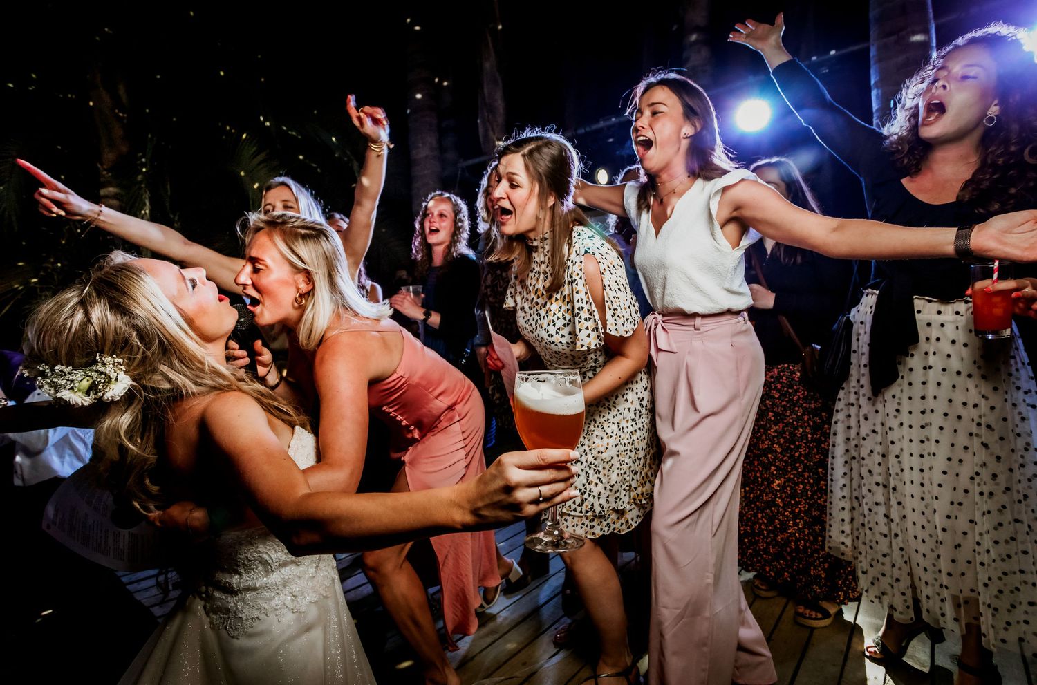 Bride and bridesmaids dancing and singing joyfully at outdoor wedding reception under warm lights.