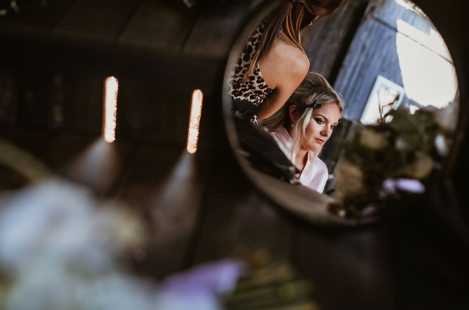 A bride getting ready is reflected in a round vintage mirror surrounded by soft floral decorations.