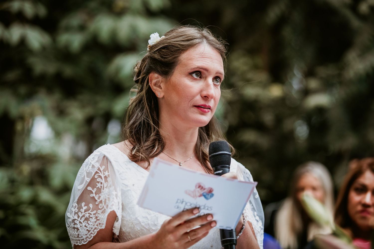 Emotional bride in lace dress reads vows into a microphone at her wedding.