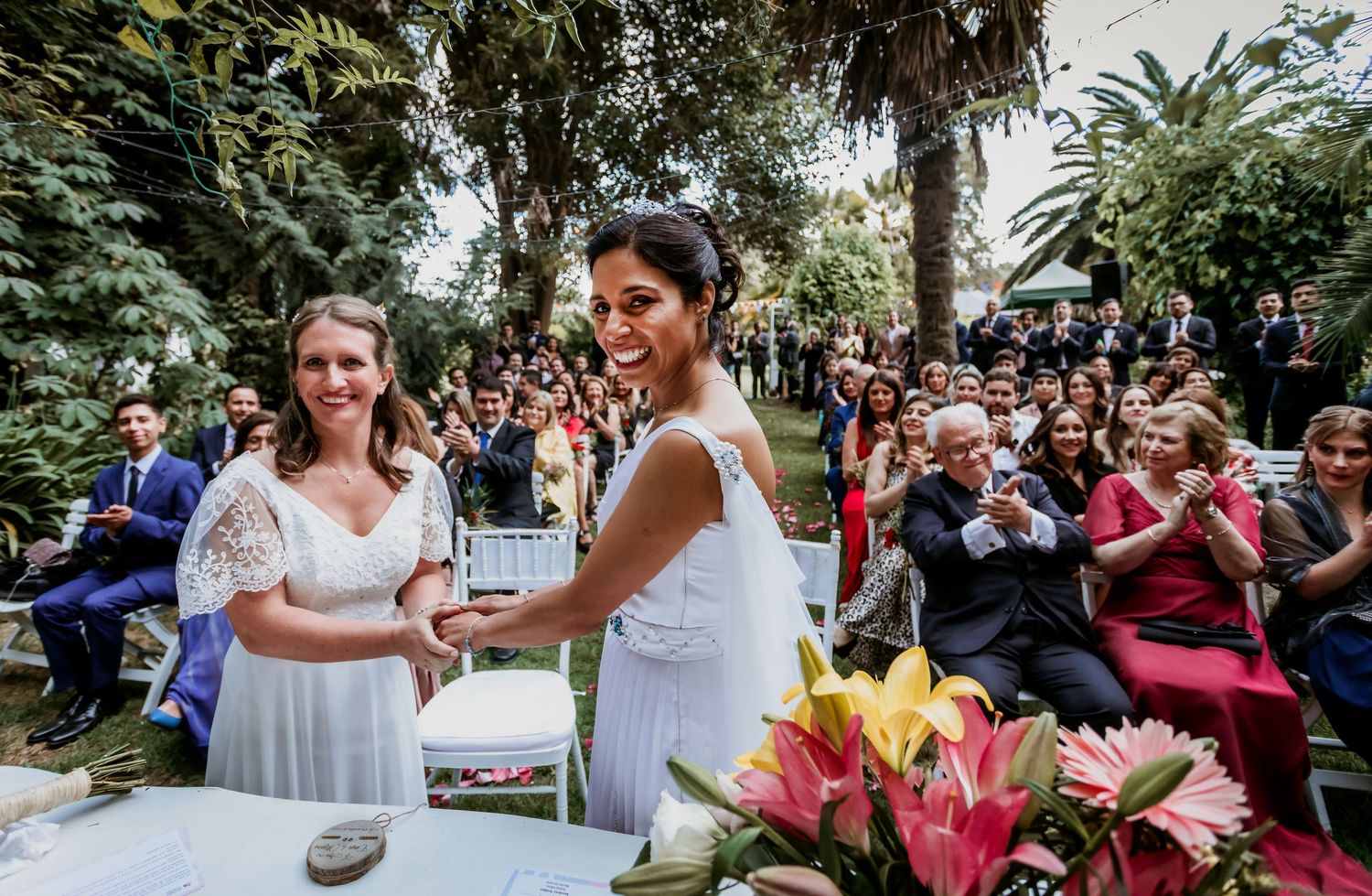 Two brides smiling and holding hands during an outdoor wedding ceremony with guests applauding in background.