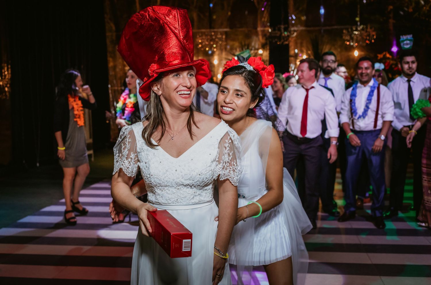 Two brides pose together smiling, one wearing a festive red hat, at a lively wedding reception celebration.