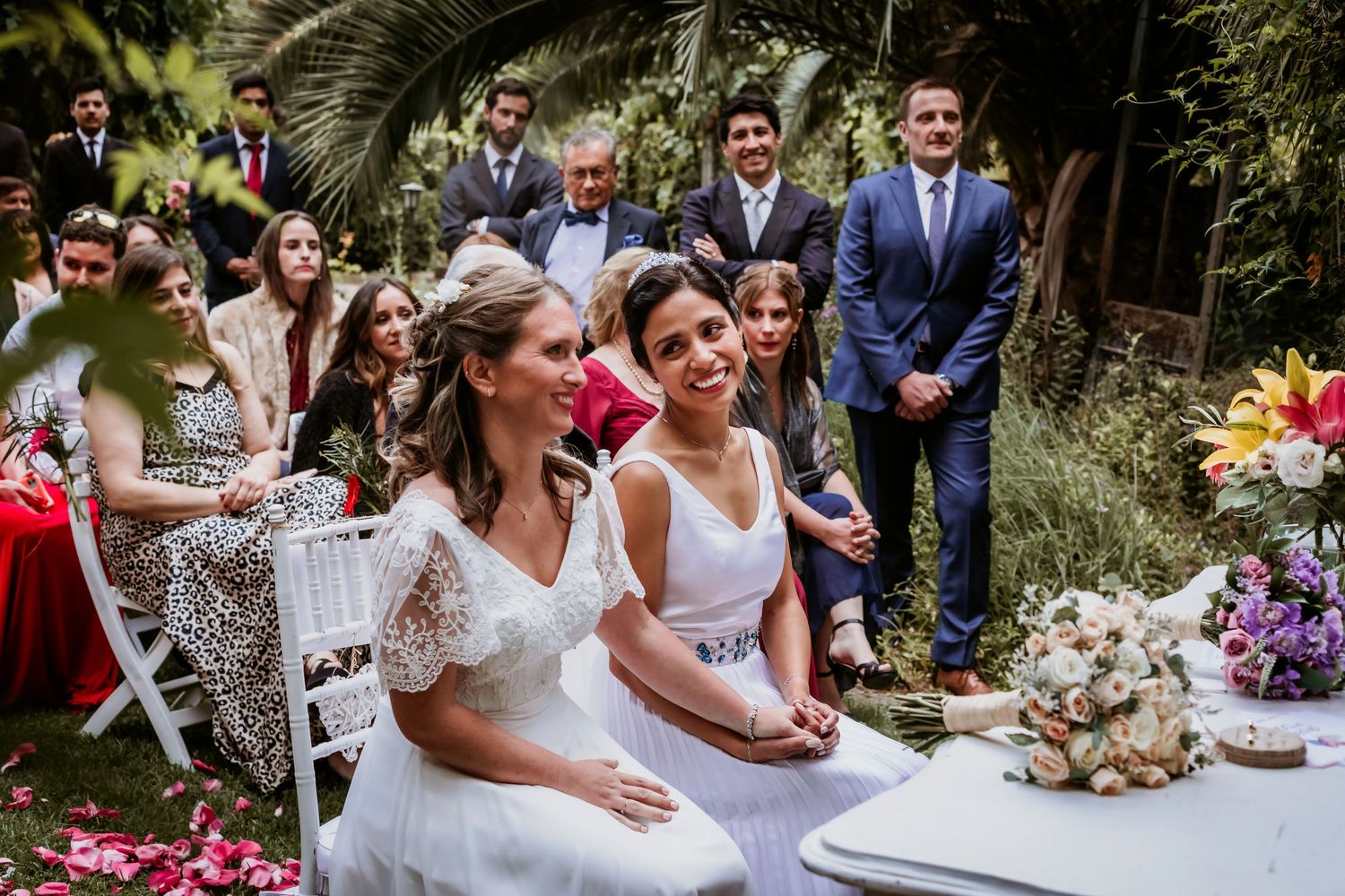 Two brides smile together at outdoor wedding ceremony table surrounded by guests and flowers.
