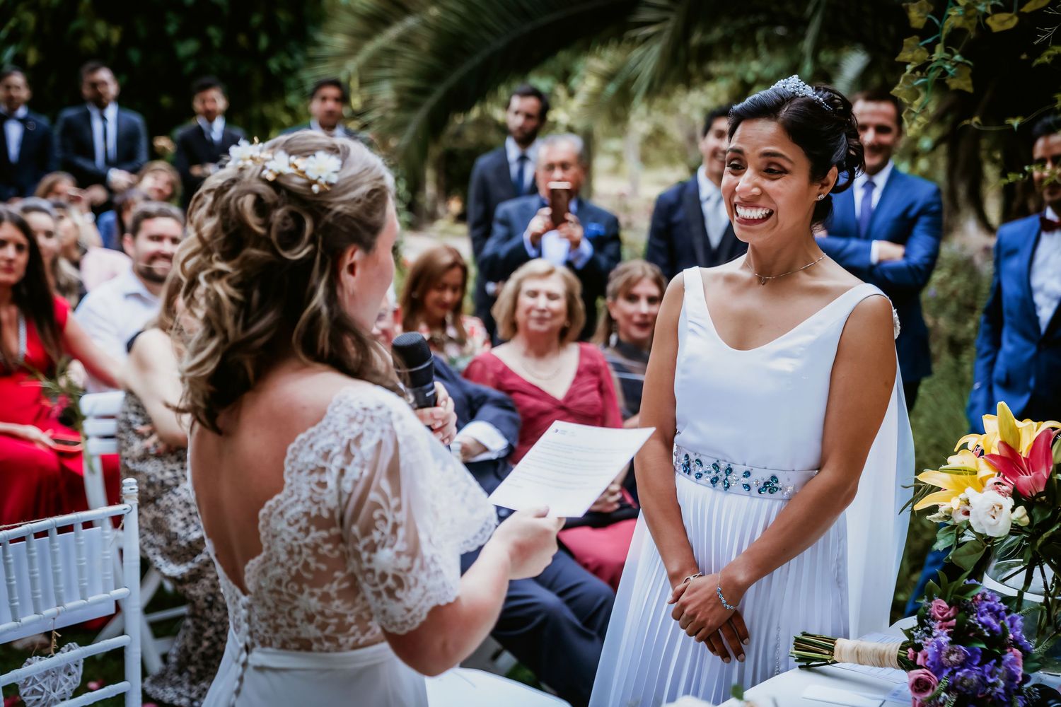 Two brides smile at each other while exchanging wedding vows at an outdoor ceremony.