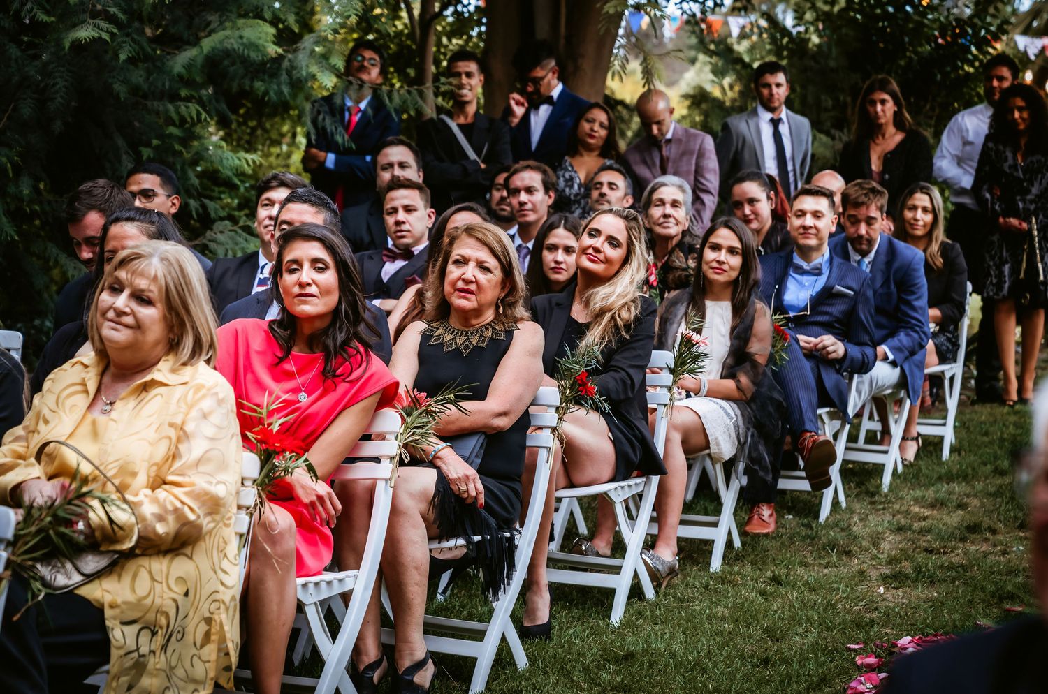 Wedding guests seated in white chairs smile and watch outdoor garden ceremony with roses and greenery backdrop.