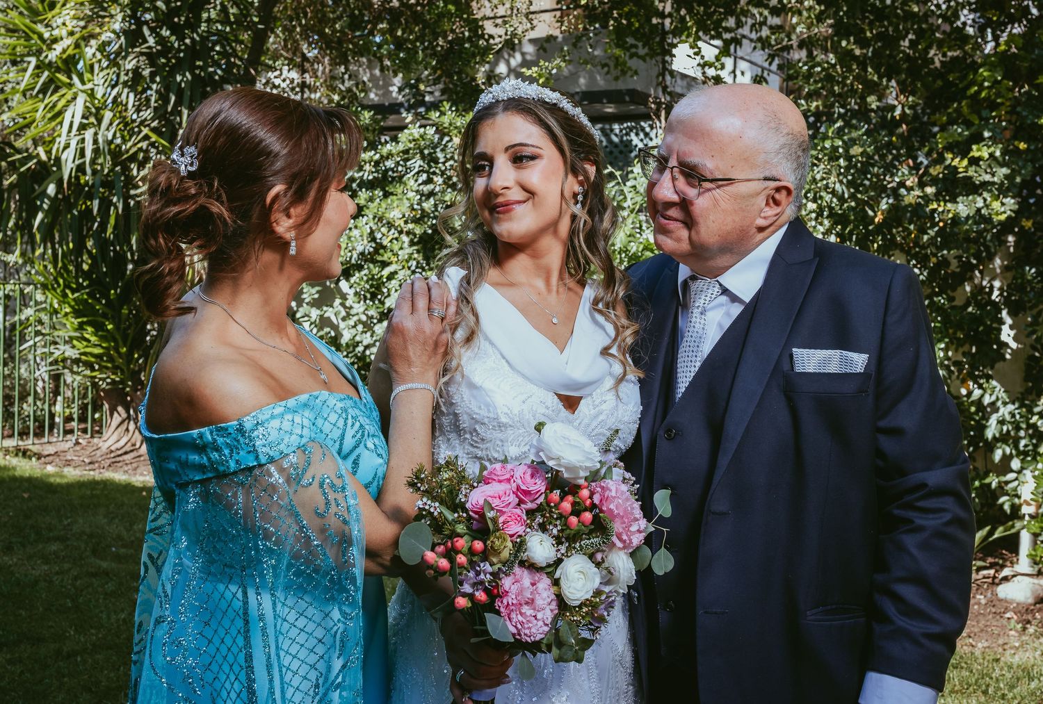 Bride in a white gown holding a pink bouquet stands with a woman in blue and an older man.