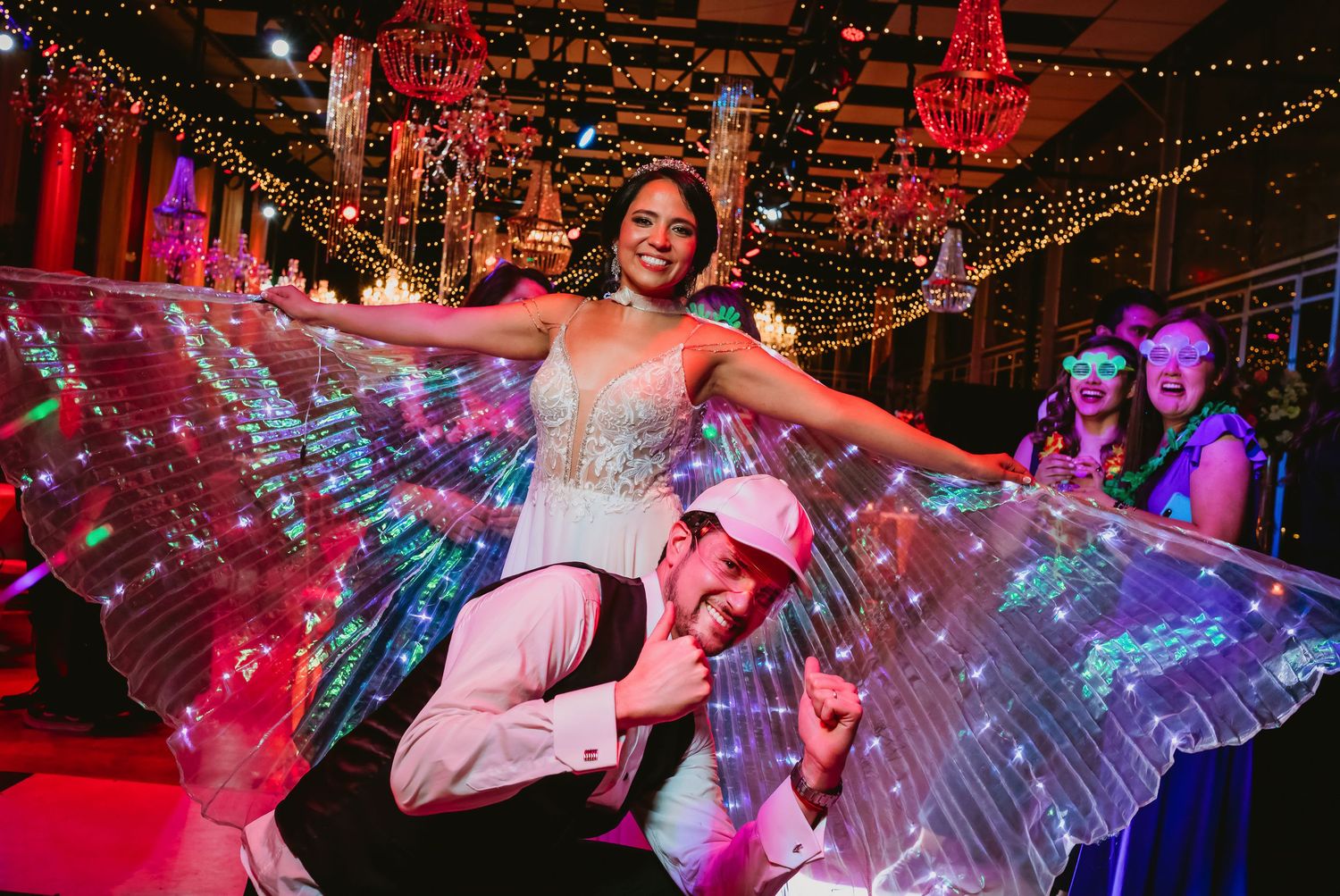 Bride in white gown with glowing LED butterfly wings lifted by groom at wedding reception.