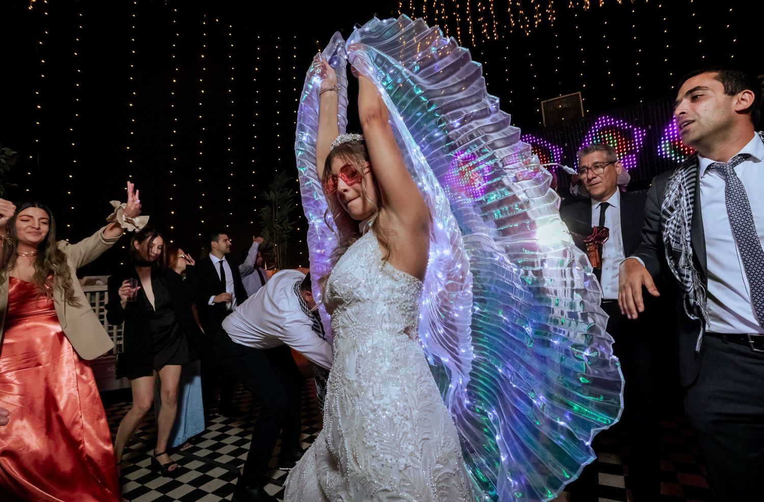Bride wearing glowing LED wings dancing joyfully with guests at a magical wedding reception party.