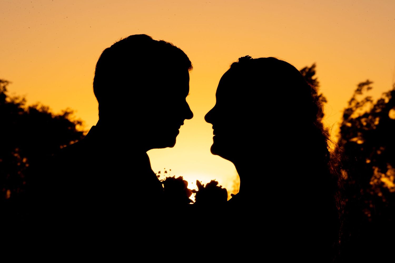 Romantic silhouette of bride and groom facing each other against a vibrant orange sunset sky.