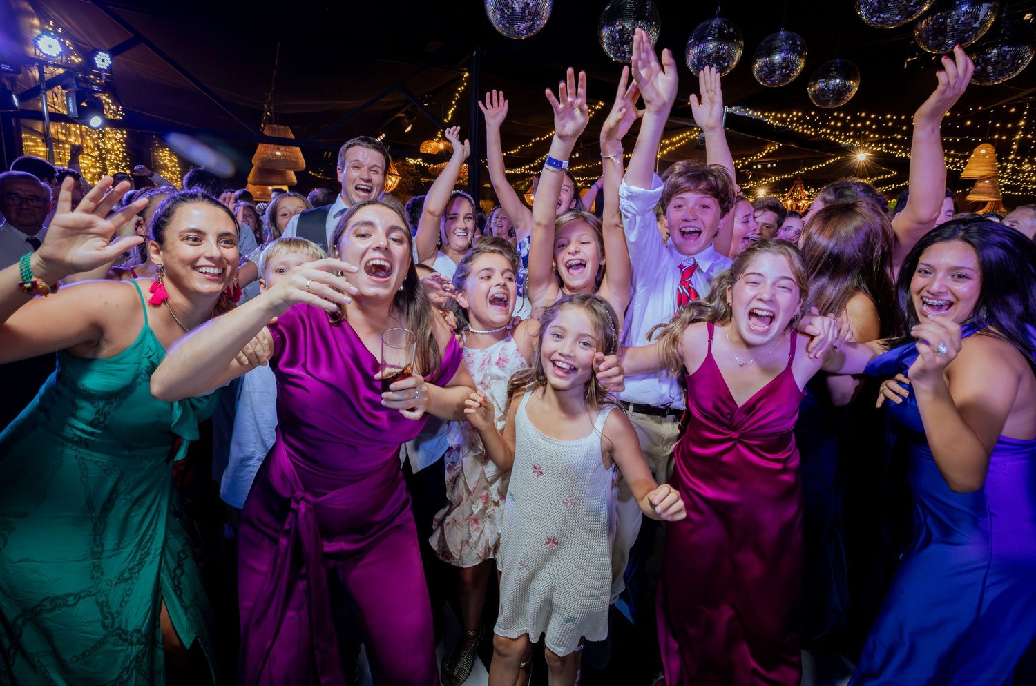 Excited wedding guests raising hands and taking selfie under disco balls at lively reception.