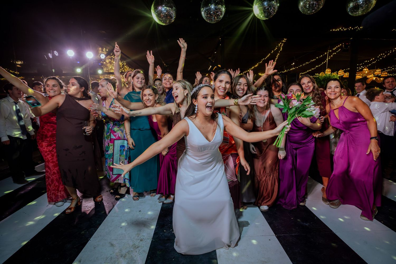 Bride tosses bouquet to excited wedding guests on dance floor with disco balls overhead.