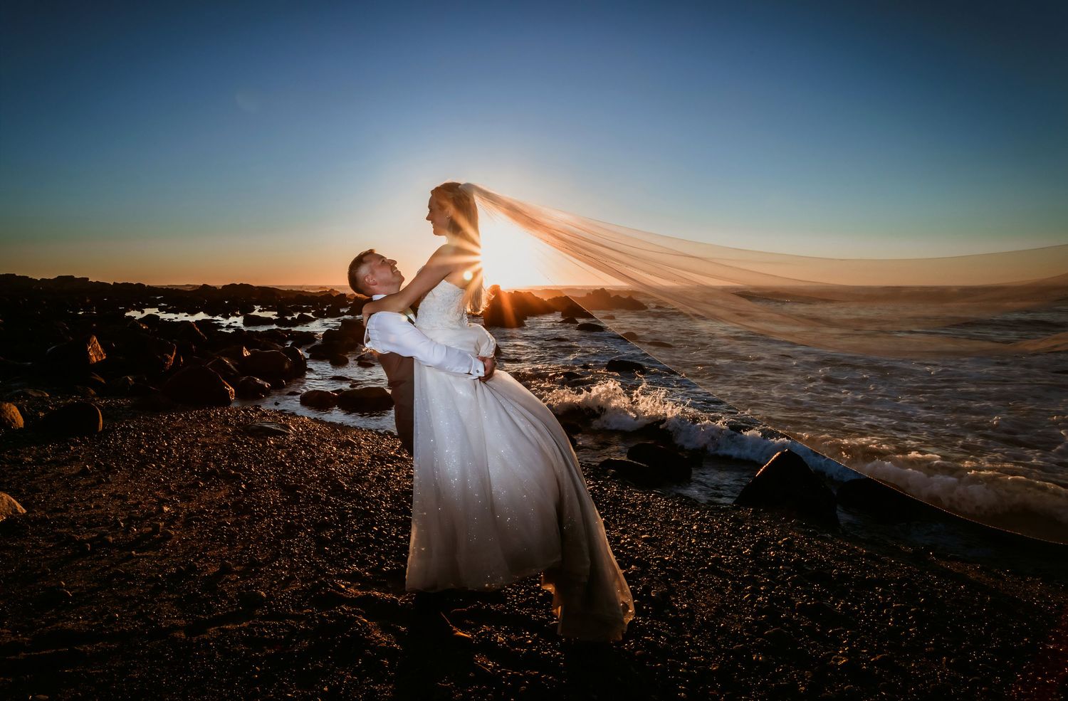 Groom lifting bride on beach at sunset, her long veil billowing in the golden light.