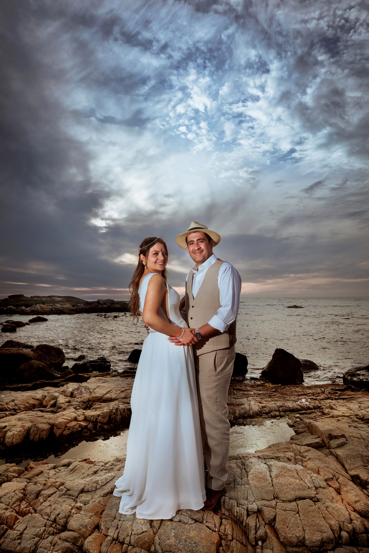 Smiling bride and groom holding hands on rocky ocean shore under dramatic cloudy sky.