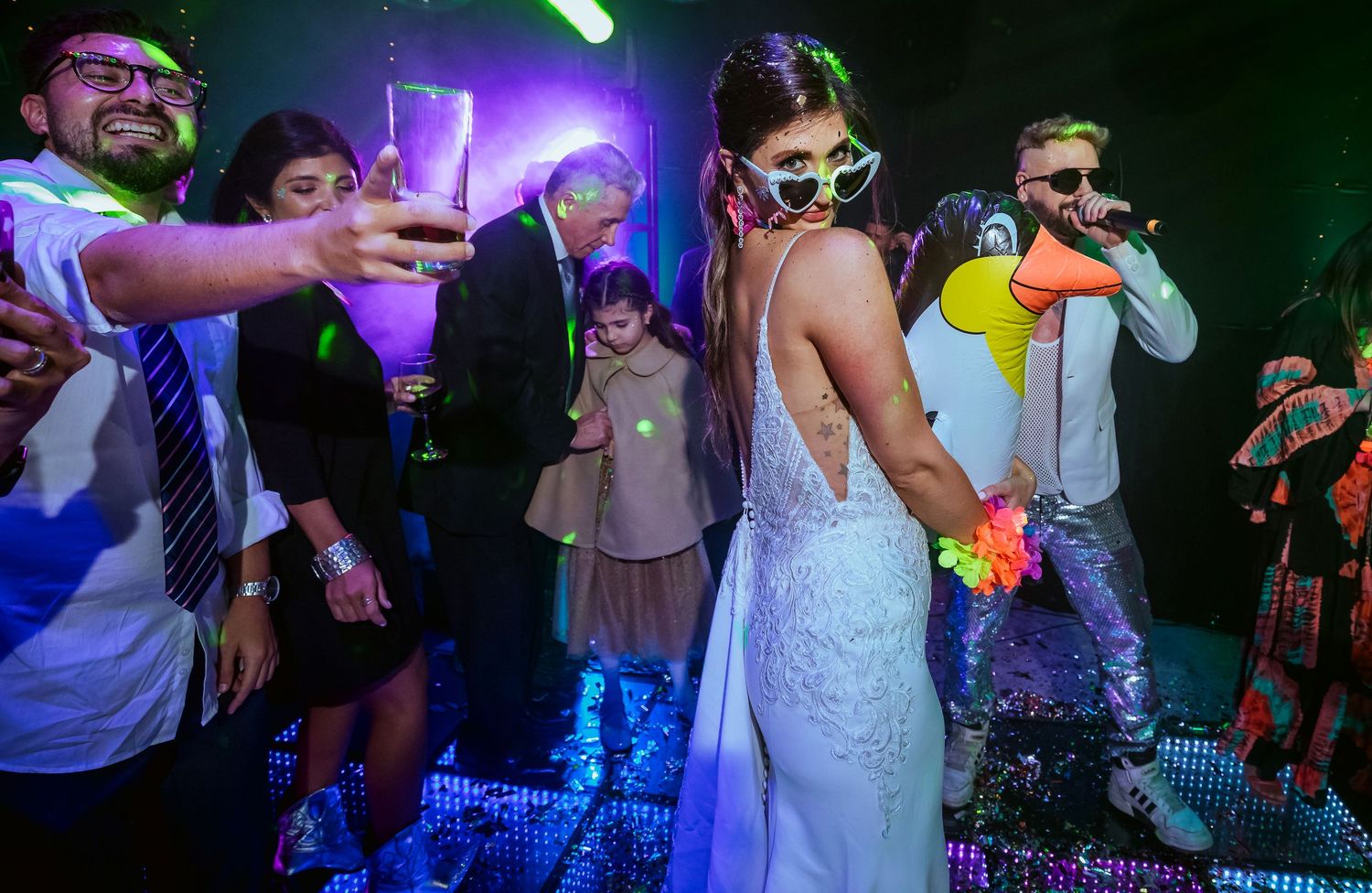 Bride in white backless gown dancing with guests at a lively wedding reception with colorful lights.