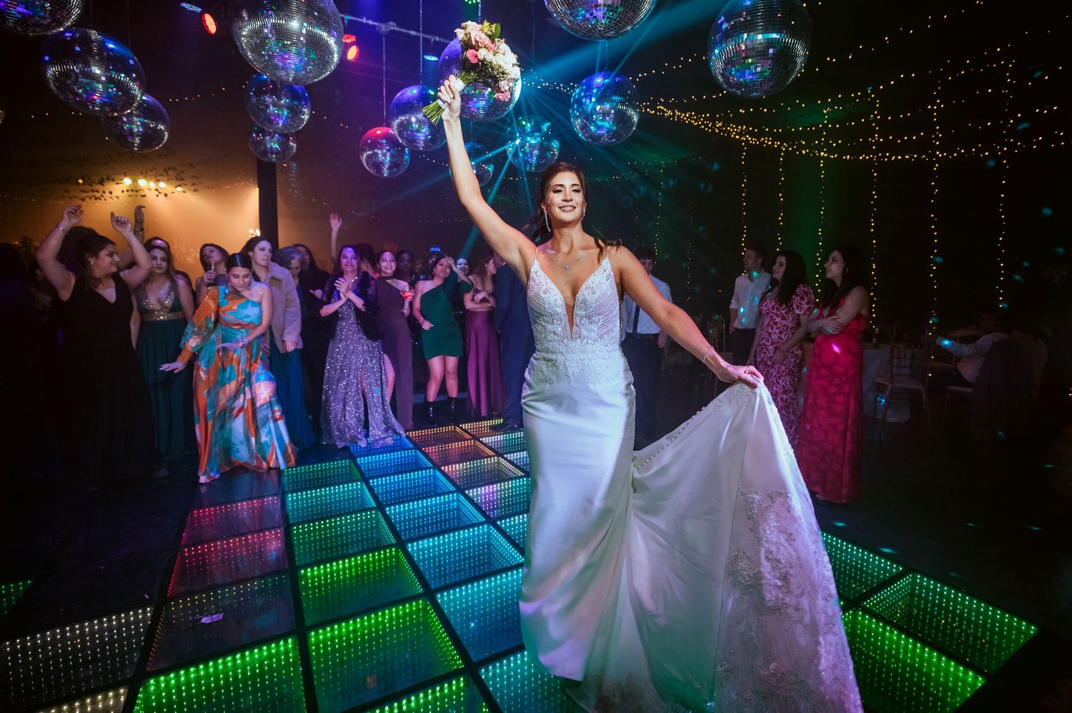 Bride holding bouquet aloft on glowing LED dance floor surrounded by guests at colorful wedding reception.