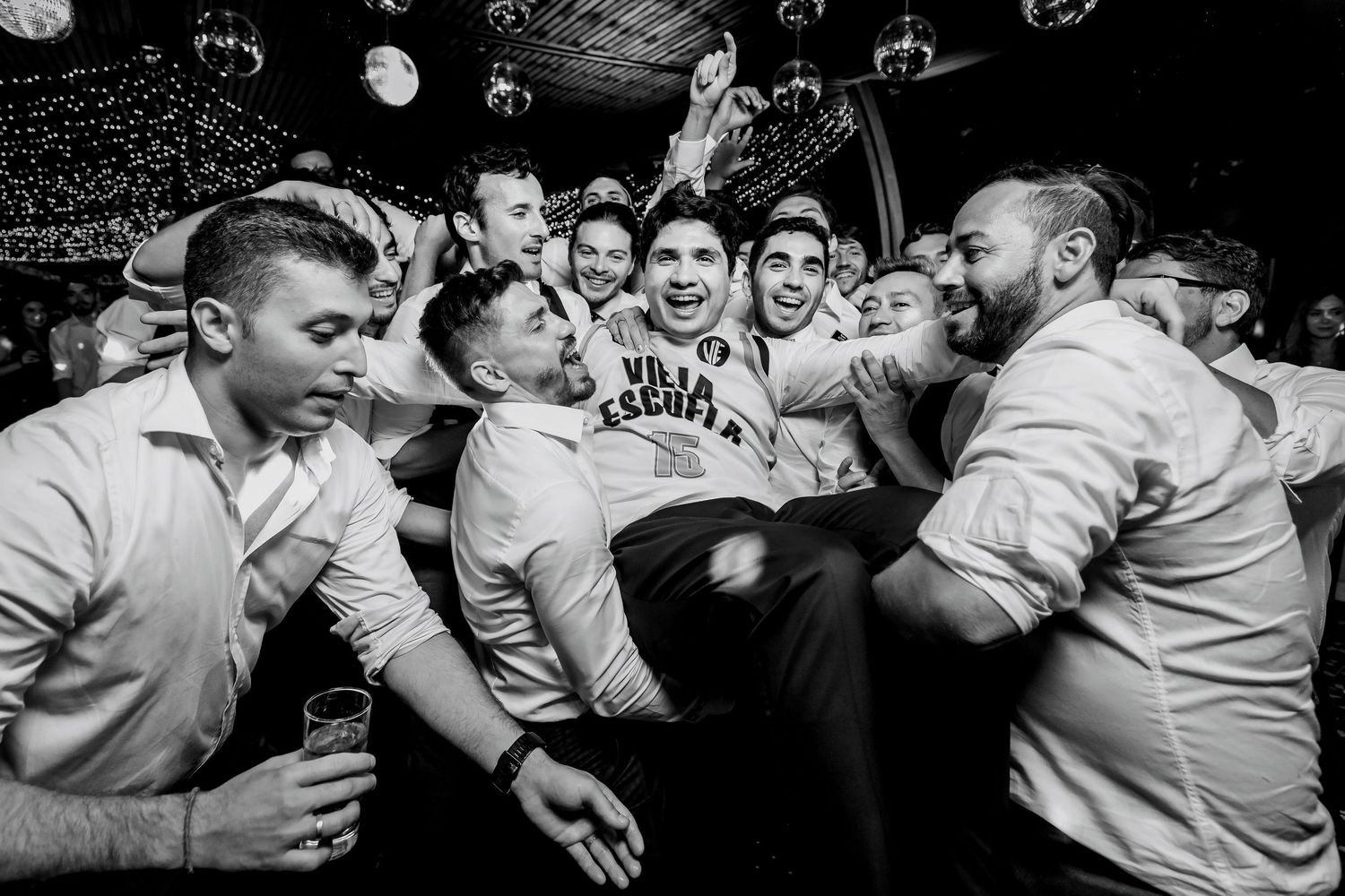 Groom lifted by groomsmen in black and white wedding party celebration photo.