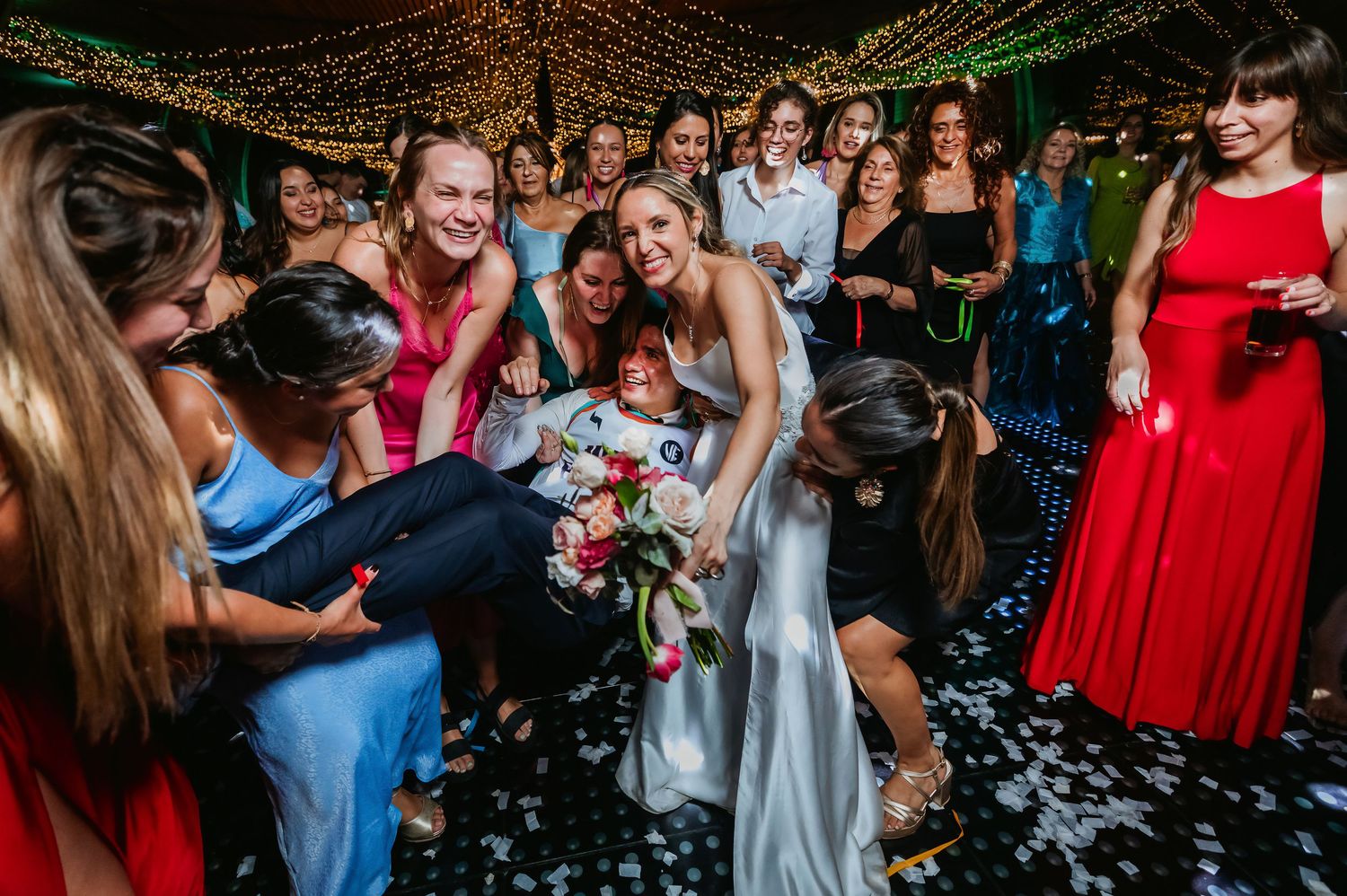 Bride in white gown surrounded by smiling wedding guests on a confetti-covered dance floor.