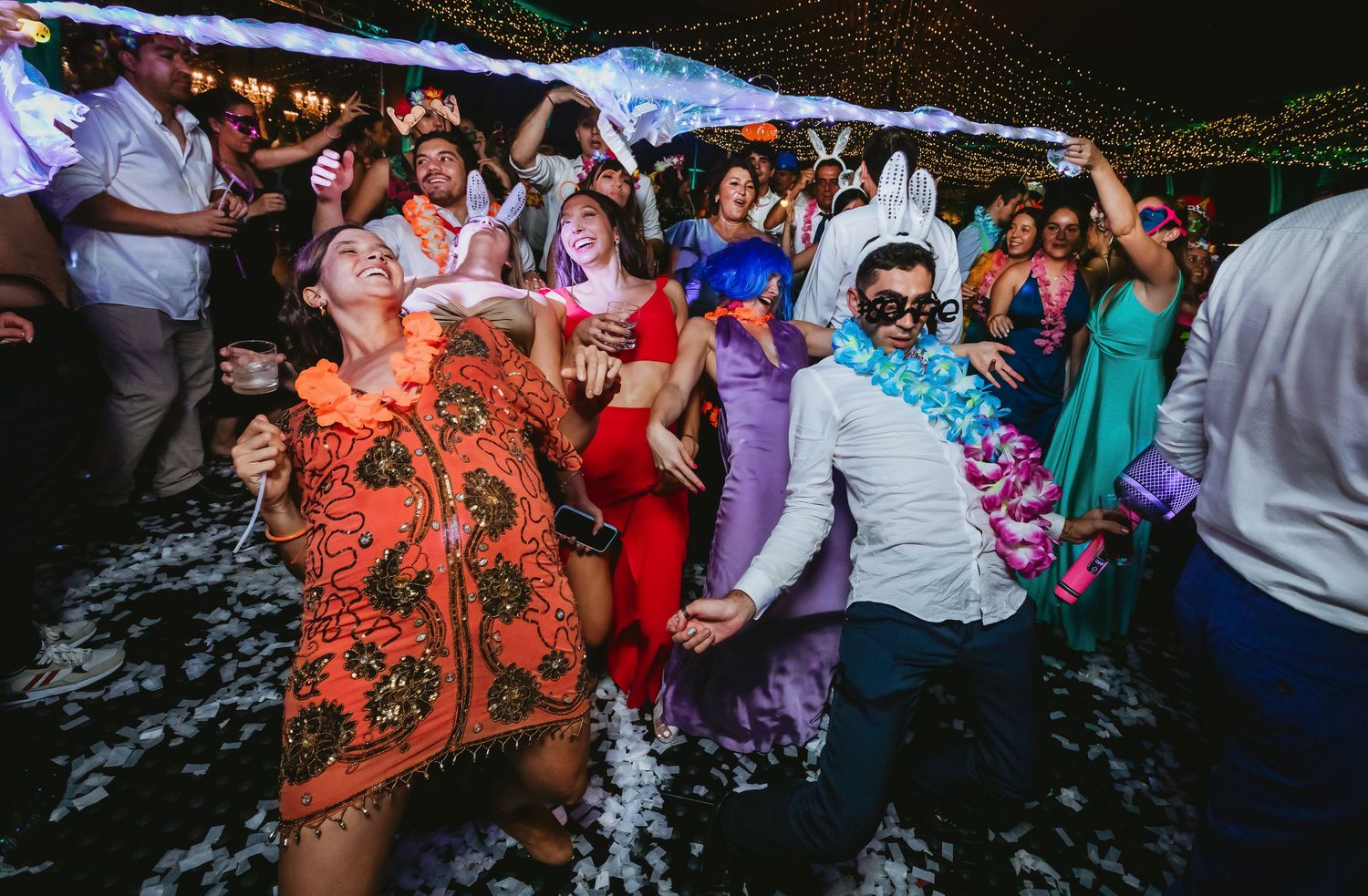 Wedding guests in colorful costumes laugh and dance together at an energetic outdoor wedding reception party.