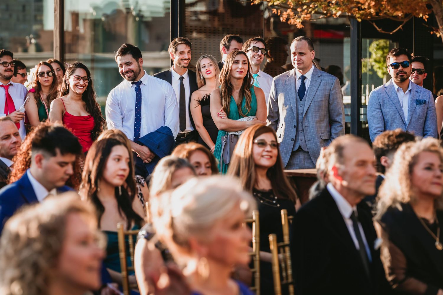Crowd of elegantly dressed wedding guests watching an outdoor ceremony attentively.