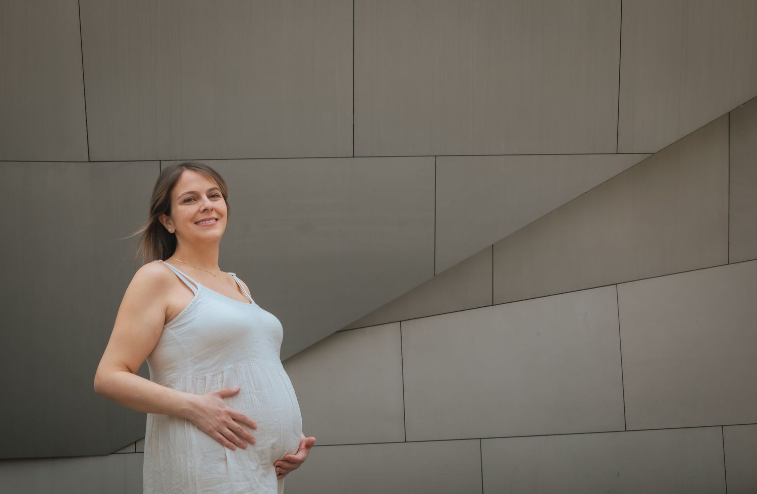 Happy pregnant woman in a white dress smiling and holding her belly against a modern gray architectural background.