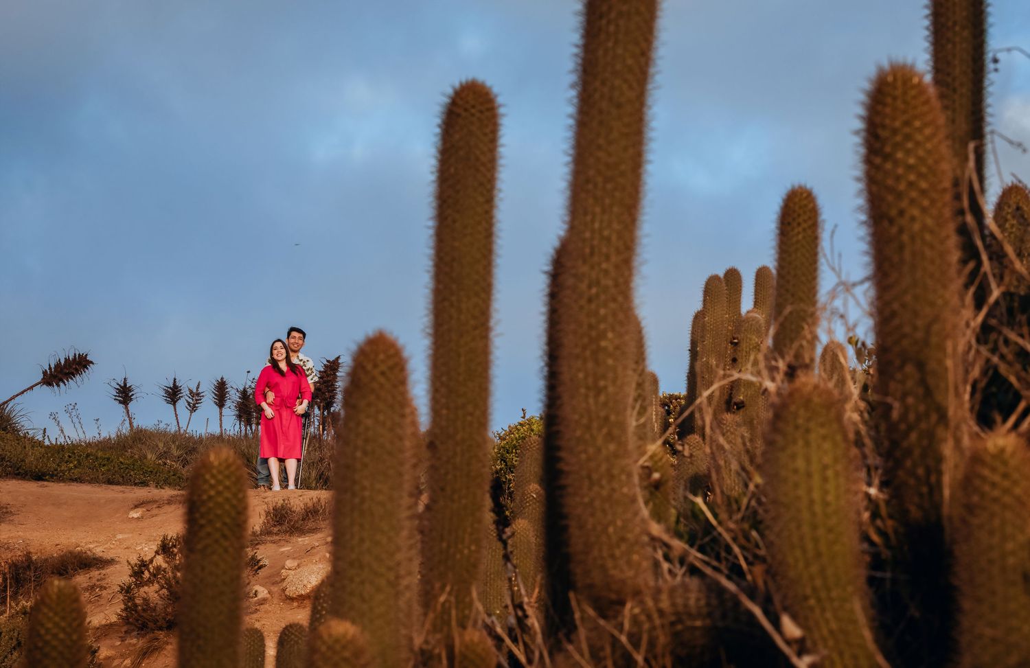 Two people in red outfits stand among tall cacti in a desert landscape under a dramatic blue sky.