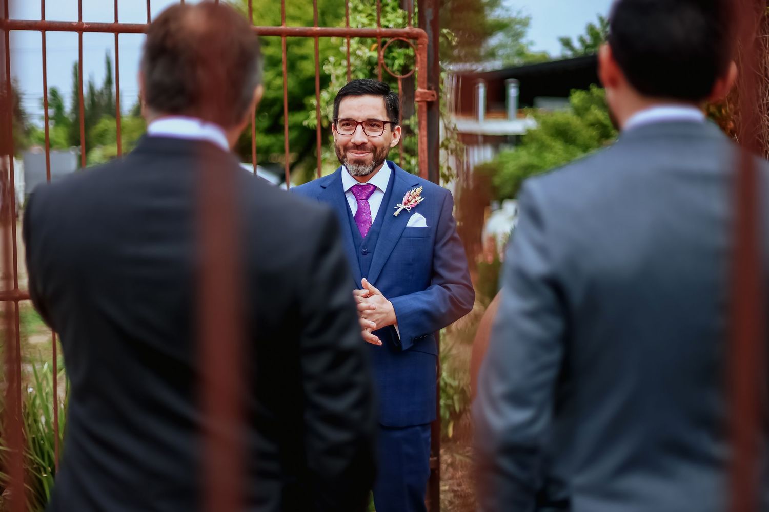 Smiling groom in blue suit greeting guests at rustic gate during outdoor wedding ceremony.