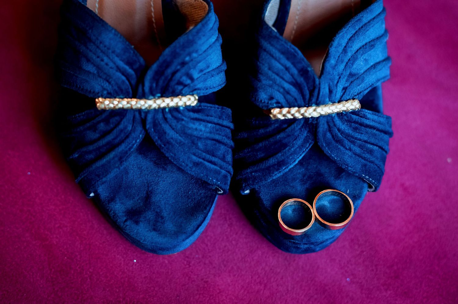 Blue suede wedding shoes with rhinestone clips beside two gold wedding bands on pink background.