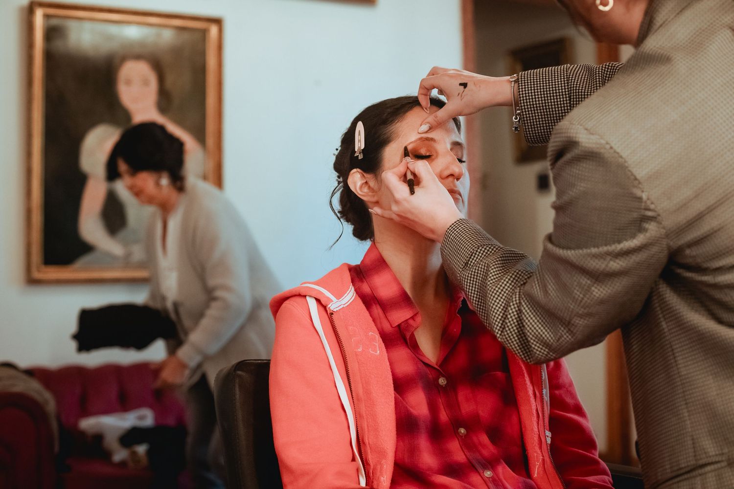 Bride getting makeup applied in red jacket while another person assists in background.