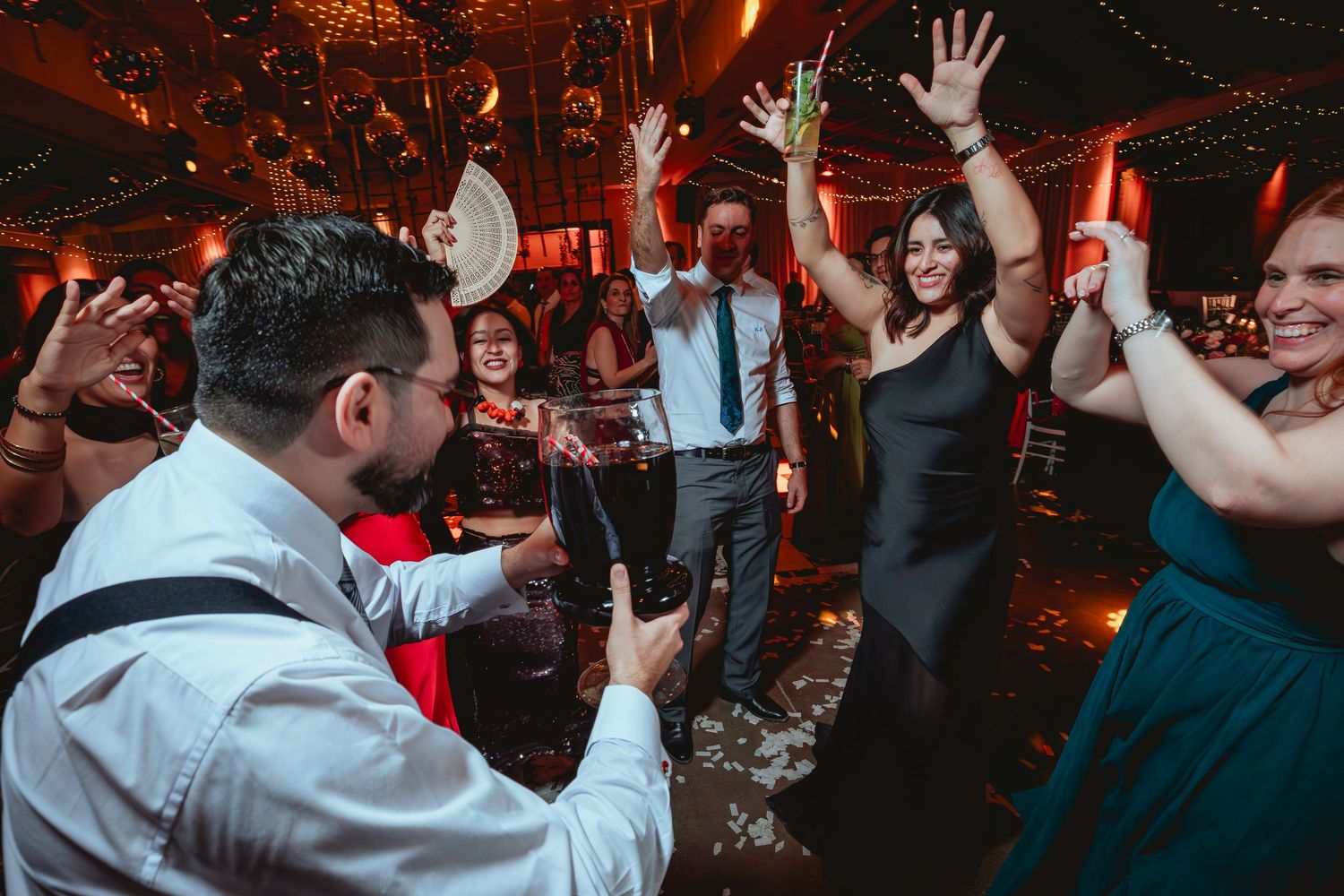 Energetic wedding guests dance and cheer at vibrant indoor wedding reception party.