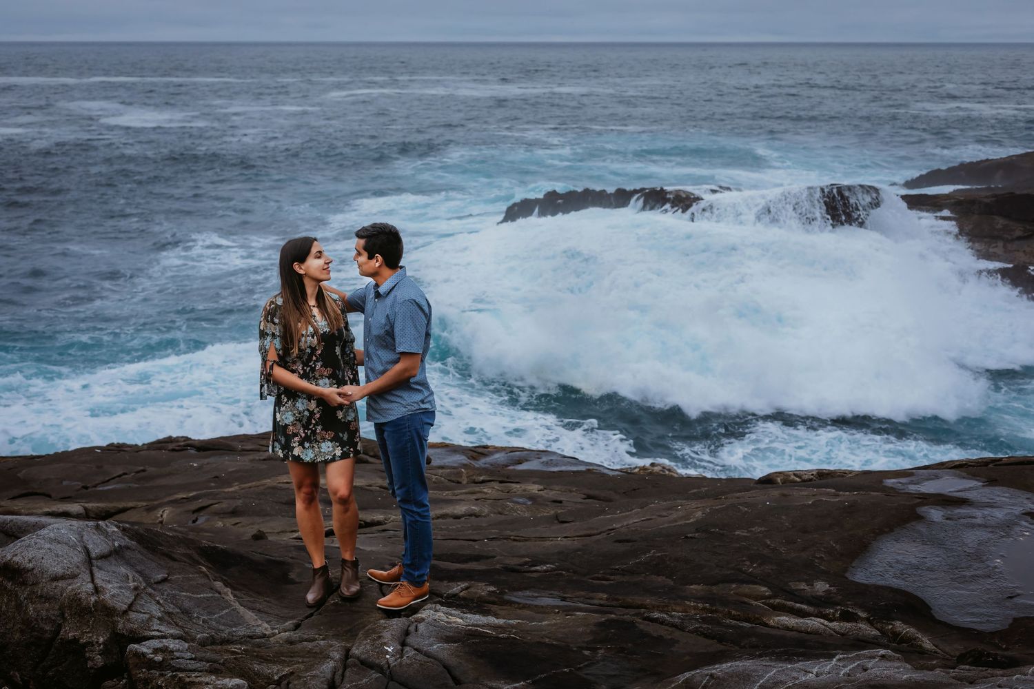 Couple standing hand in hand on rocky shore with massive ocean waves crashing behind them.