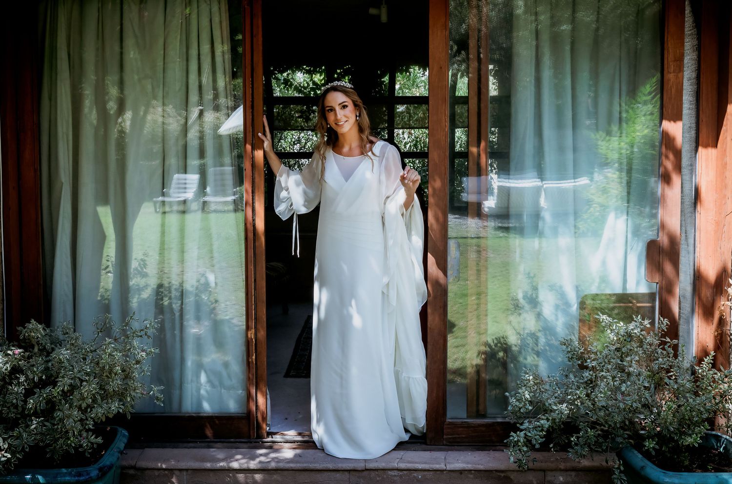 Bride in a long white dress with bell sleeves stands in open wooden doors surrounded by greenery.