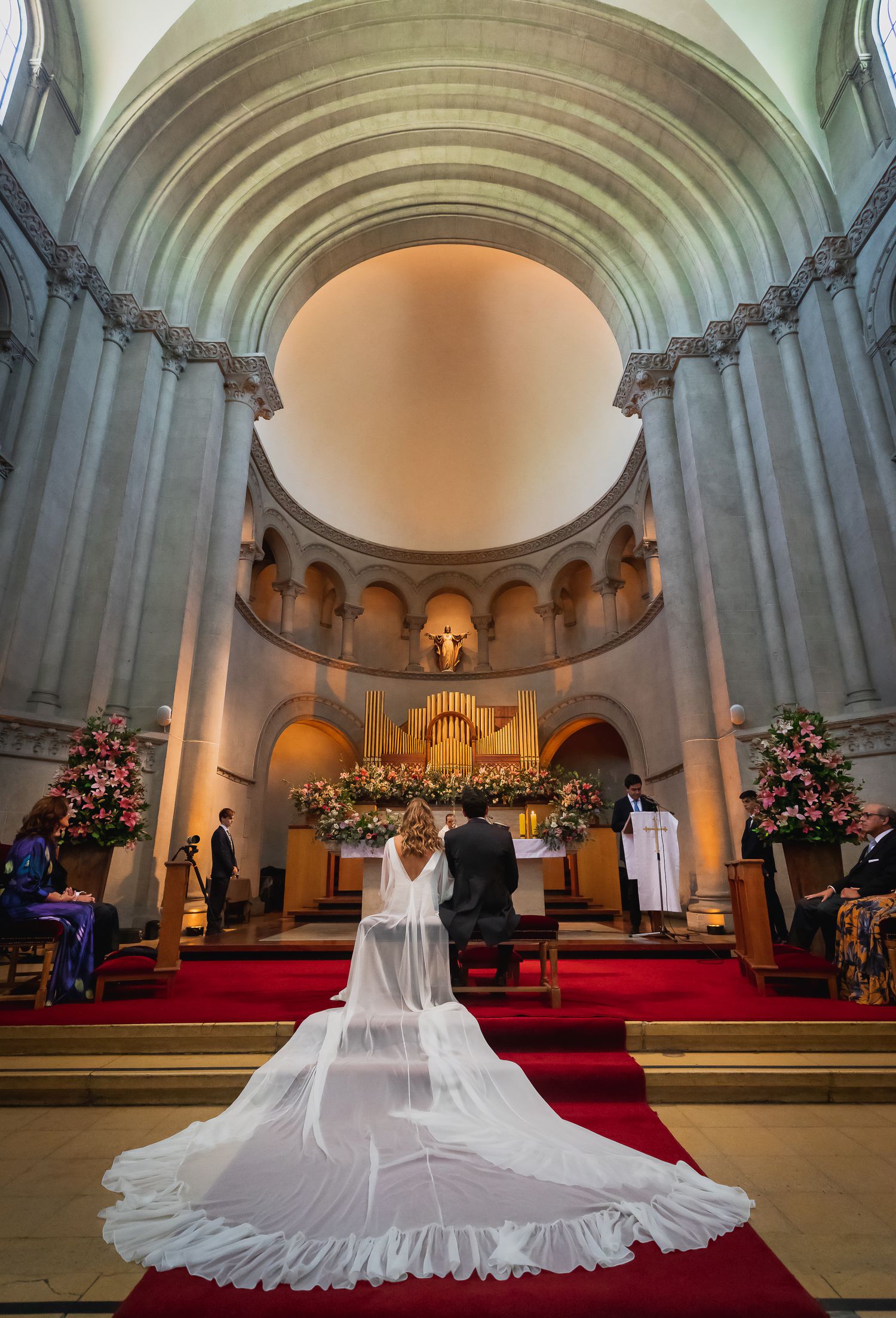 Bride in white dress with flowing veil approaching ornate church altar during ceremony.