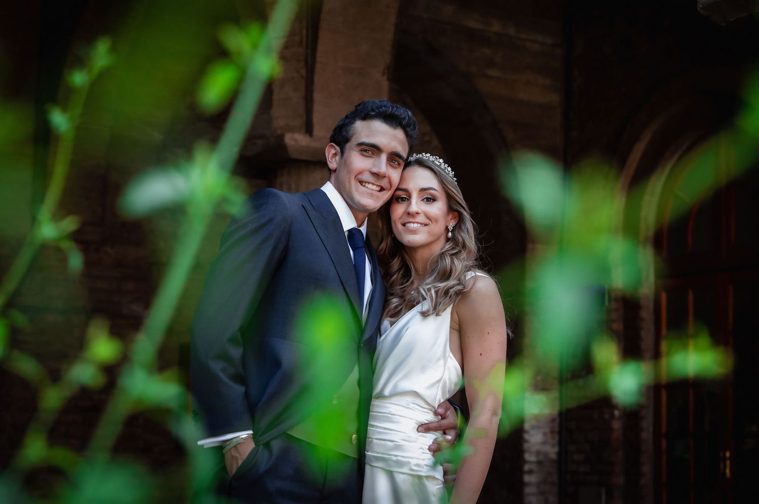 Bride and groom smiling together outdoors surrounded by green foliage near a rustic brick building.