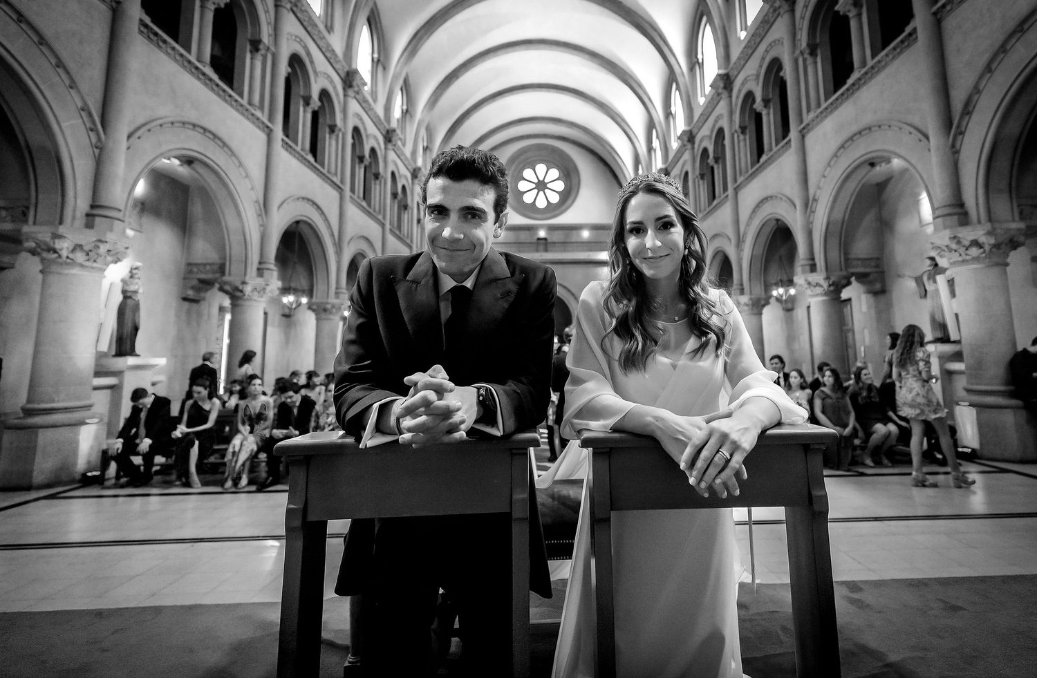 Black and white photo of smiling bride and groom kneeling at altar table inside a grand church.