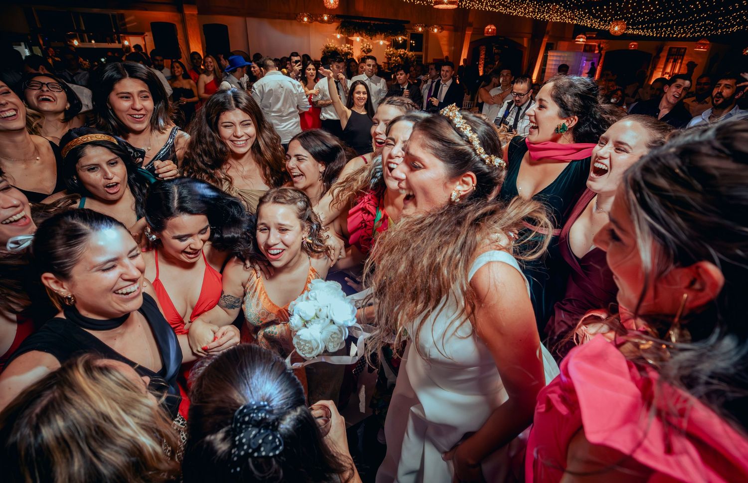 Bride surrounded by excited female guests celebrating at a lively wedding reception.