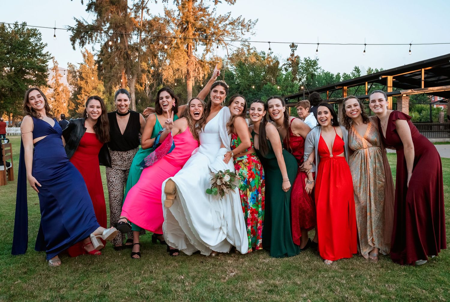 Bride poses with bridesmaids in colorful gowns outdoors at golden hour wedding celebration.