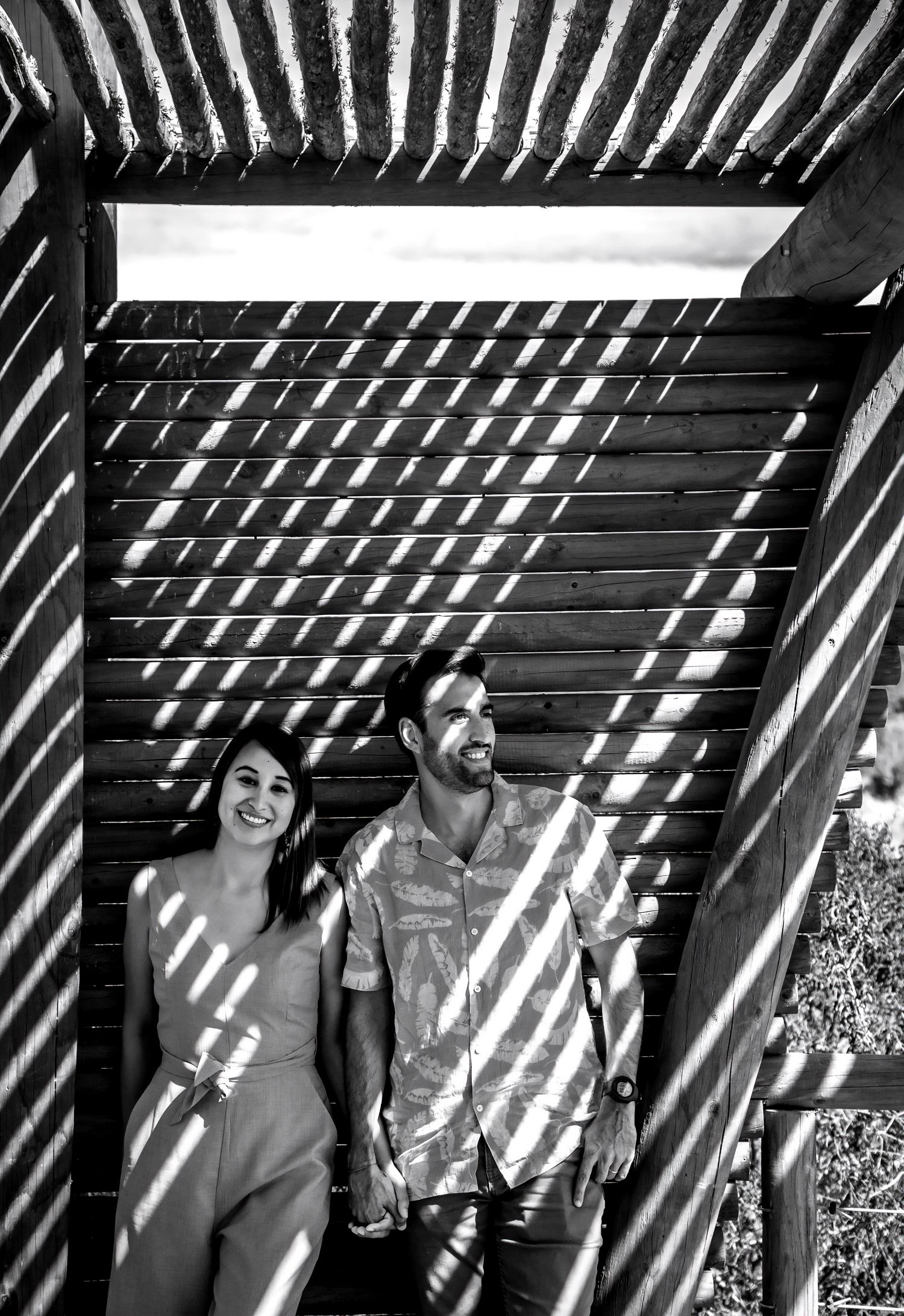 Black and white photo of a smiling couple holding hands under a striped pergola in sunlight.