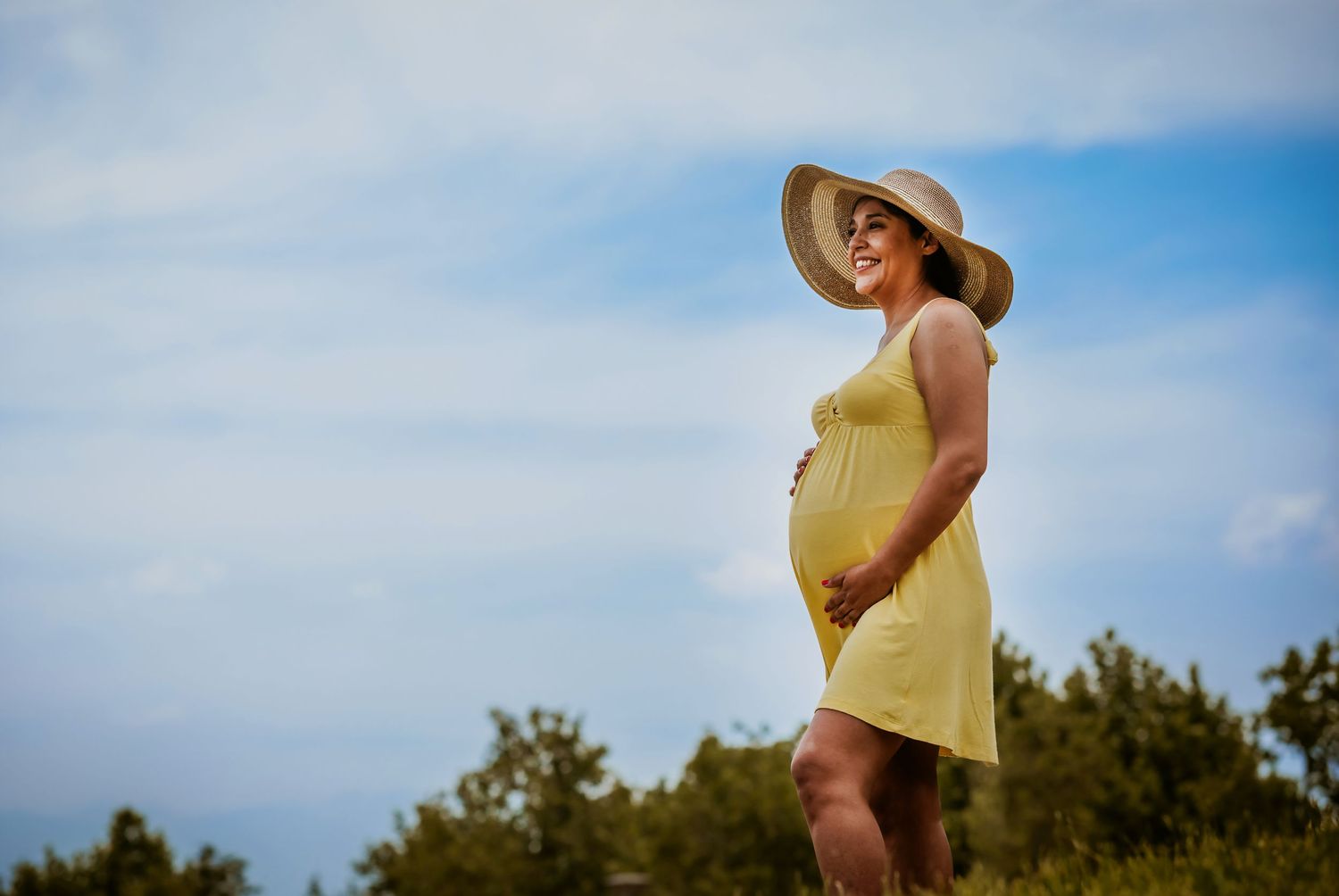 Pregnant woman in yellow dress and sun hat standing outdoors on a sunny day, cradling her baby bump.