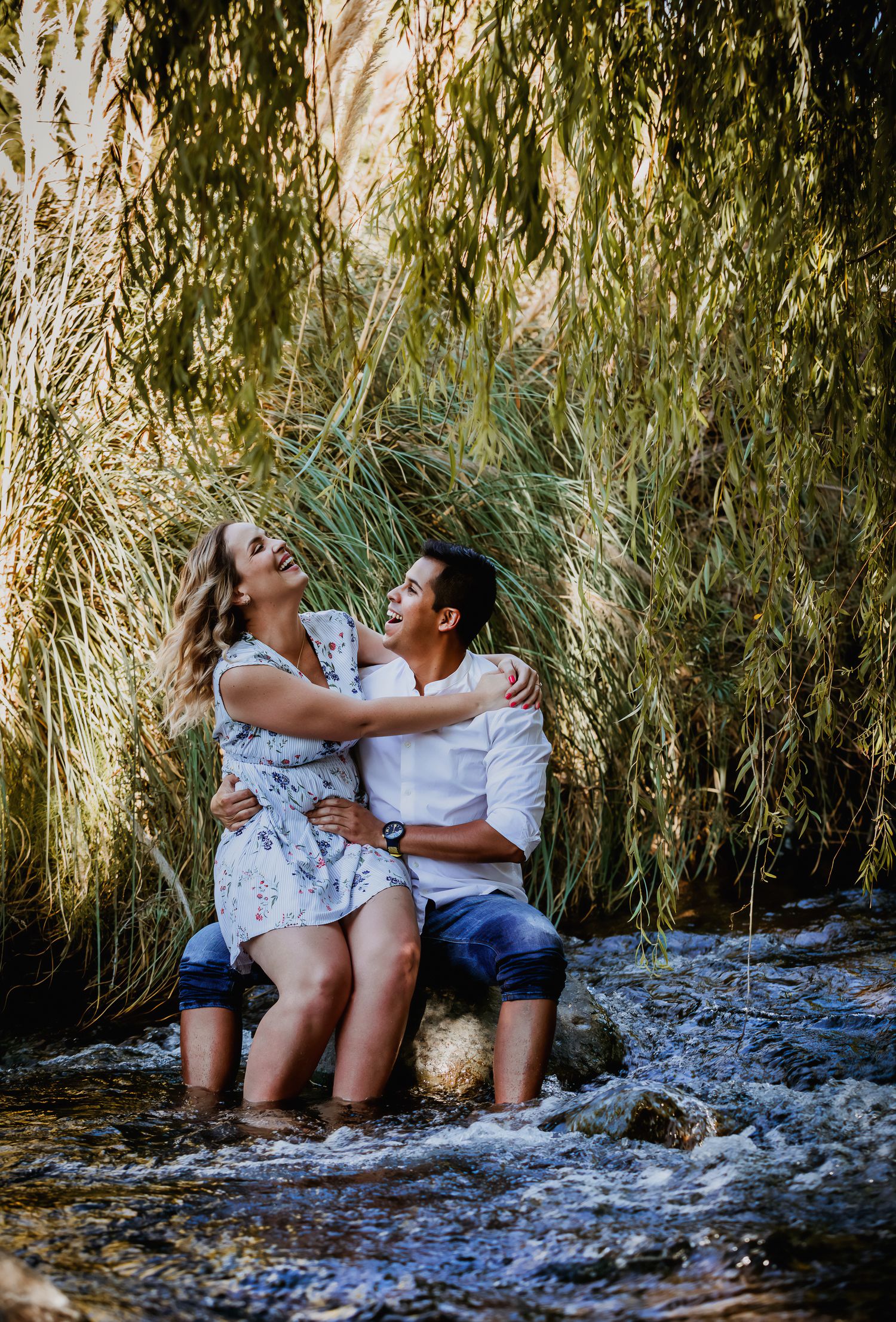 A happy couple sits embracing on rocks in a flowing stream, surrounded by lush green reeds, laughing together.