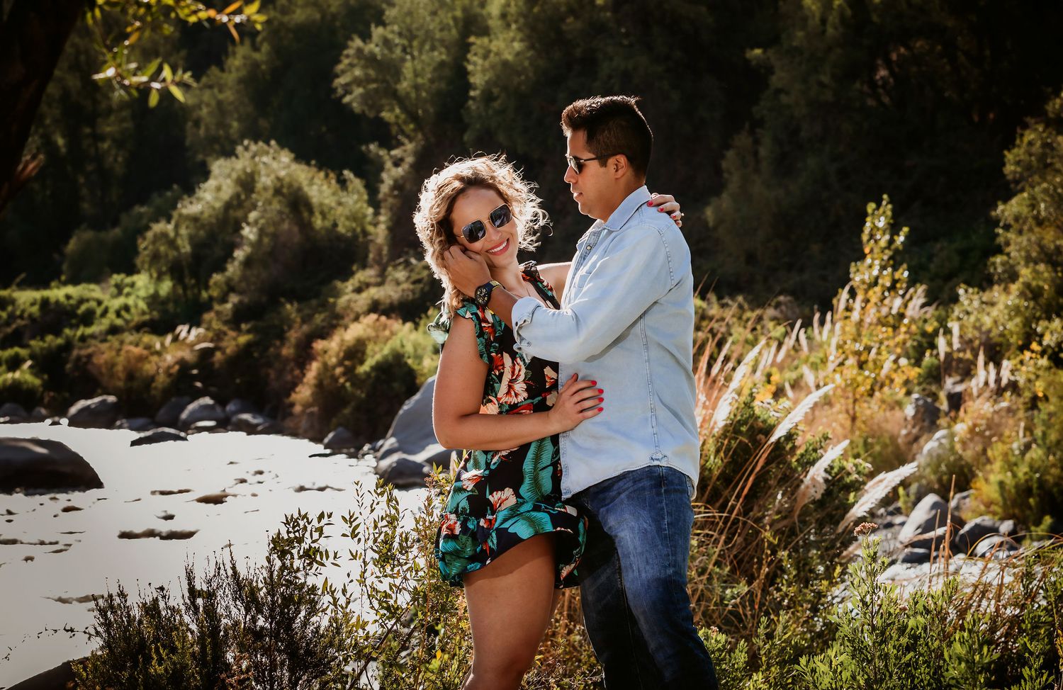 Couple embracing outdoors near a river surrounded by lush green trees and rocks on a sunny day.