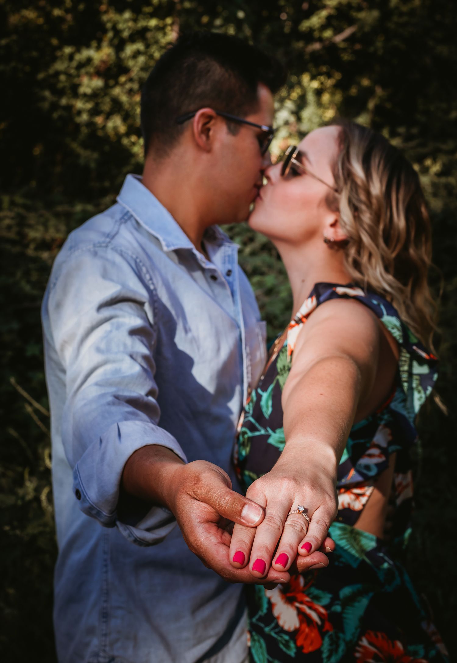 Couple sharing a romantic kiss outdoors while showing off an engagement ring, surrounded by lush green foliage.