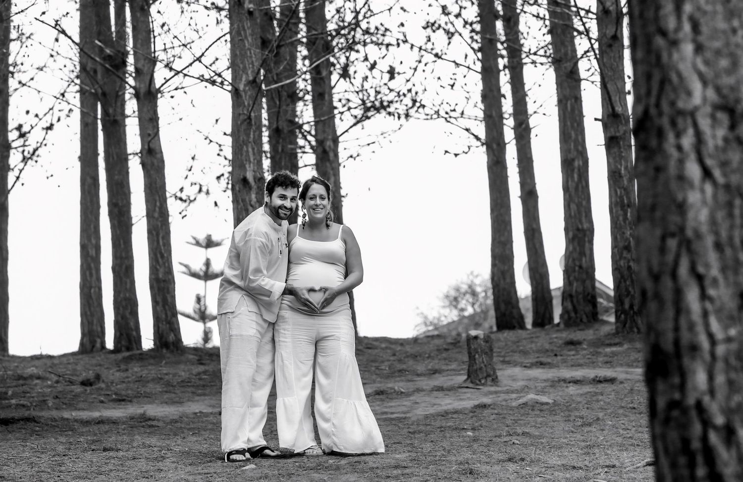 Expecting couple smiling together among tall pine trees in black and white maternity photo.