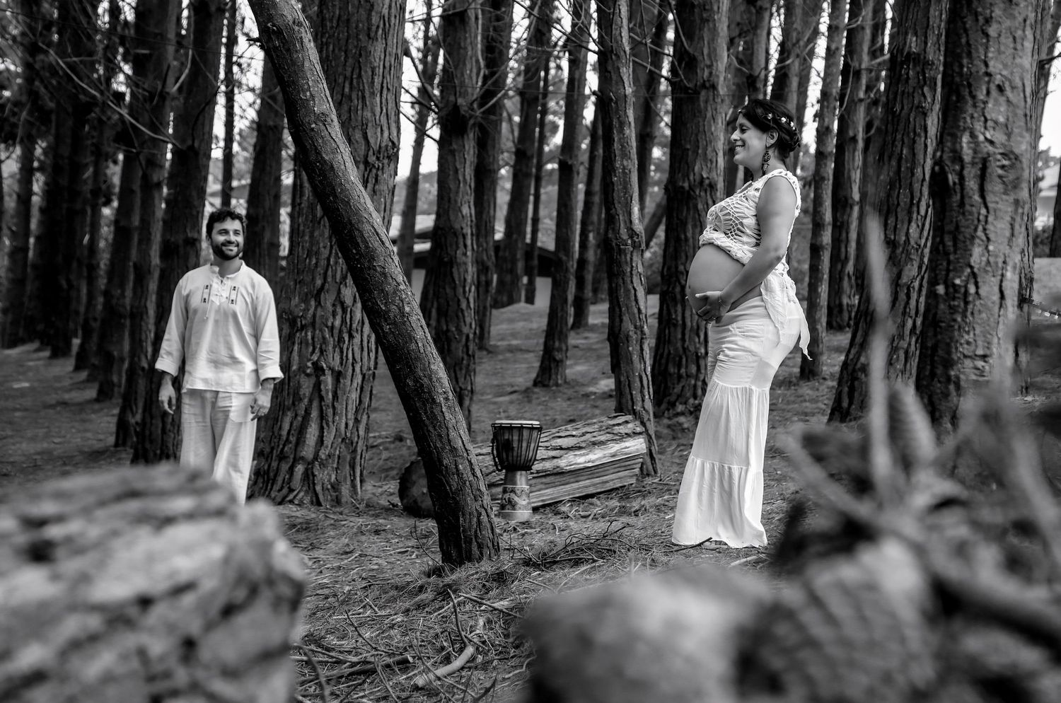 Black and white maternity photo of a pregnant woman in white dress with a man among tall pine trees in a forest.