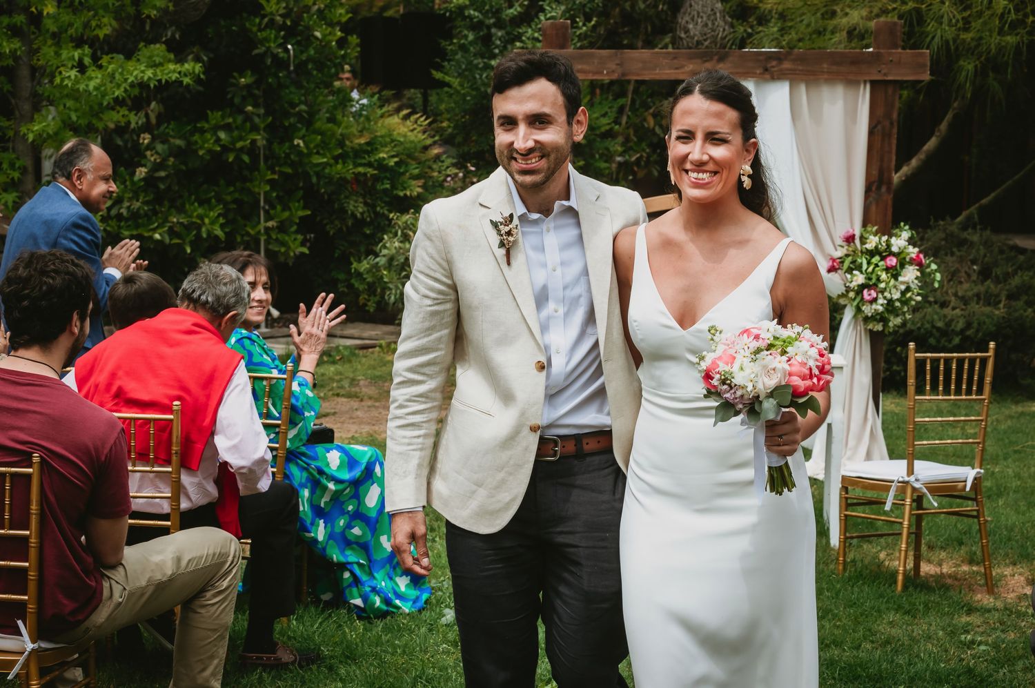 Smiling bride and groom walk down outdoor aisle after wedding ceremony, bride holding pink bouquet.