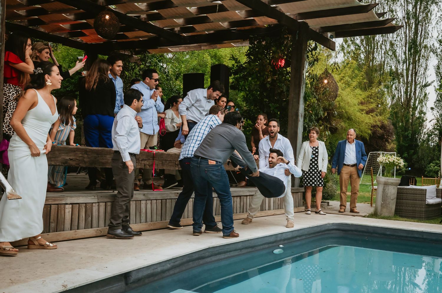 Wedding guests celebrating outdoors near a pool under a pergola, with lush greenery in the background.