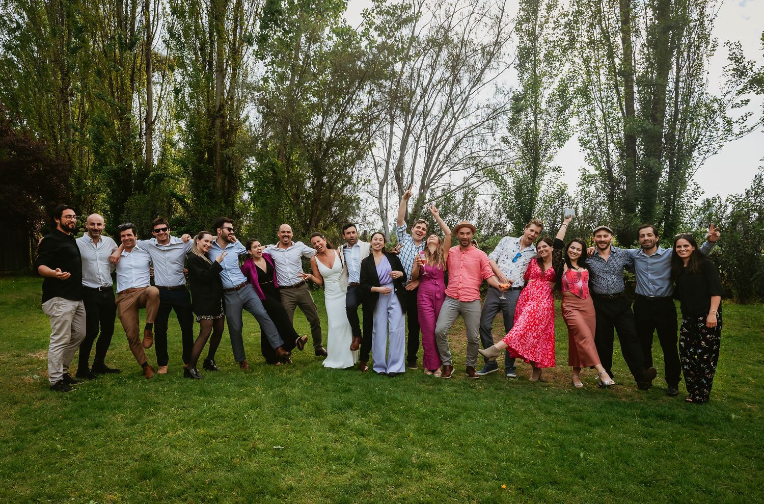 Large wedding group photo outdoors on green lawn with tall trees, guests posing joyfully with bride and groom in center.