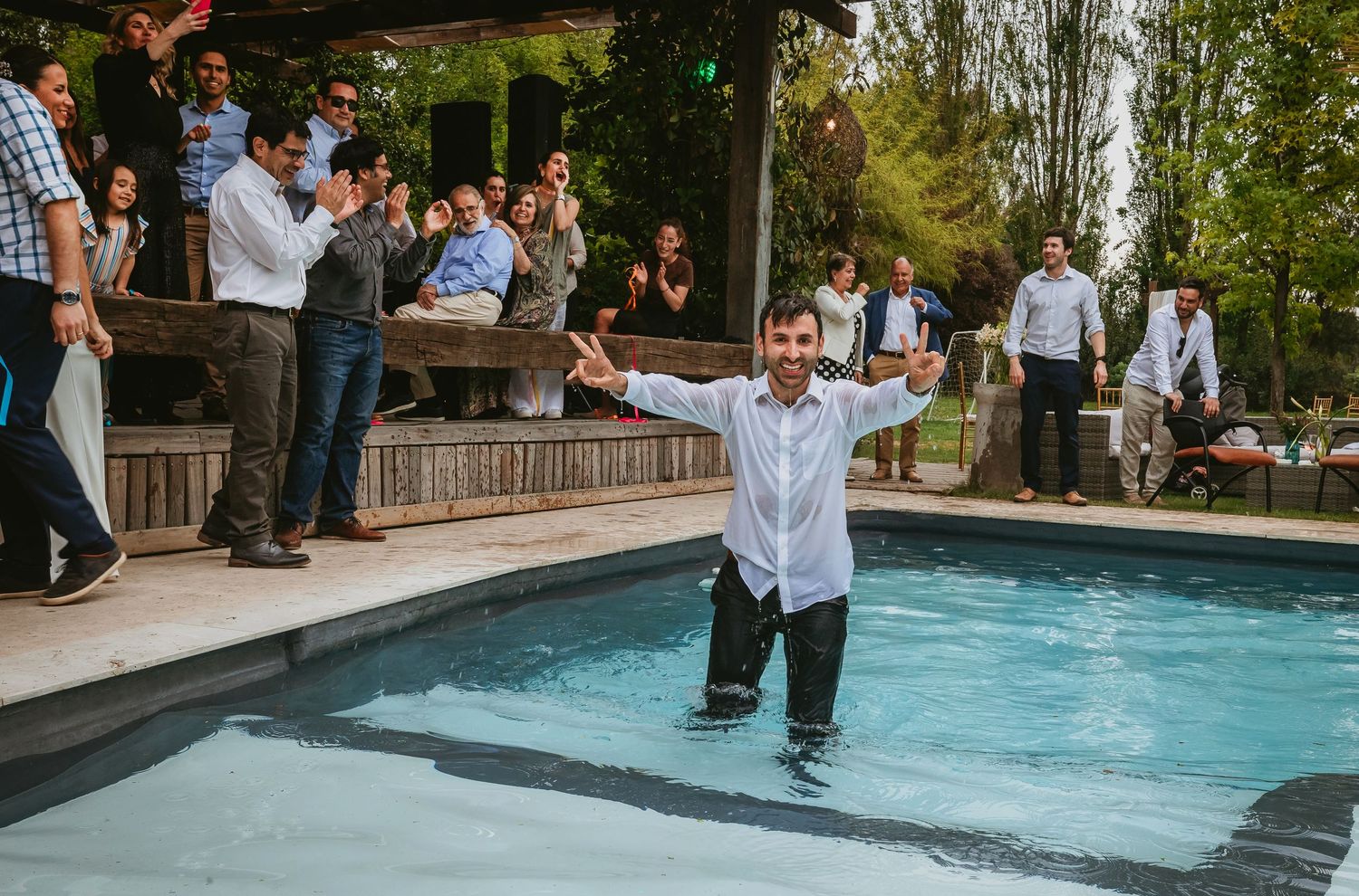 Man stands in pool fully clothed, arms raised, while guests celebrate poolside at an outdoor party.
